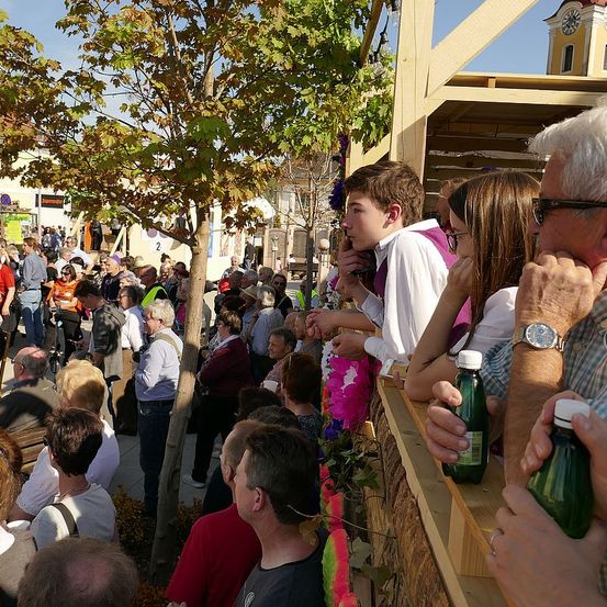 Eine Menschenmenge steht draußen und beobachtet ein Ereignis. Ein Junge in einem weißen Hemd lehnt sich an eine Holzbarriere. In der Nähe hält ein Erwachsener eine grüne Flasche. Im Hintergrund ist ein Glockenturm zu sehen.