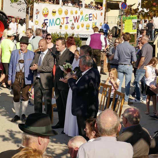 Eine Menschenmenge steht auf einer Straße, wobei einige Personen in traditioneller Kleidung gekleidet sind. Eine Person hält einen Blumenstrauß. Dahinter befindet sich ein Banner mit der Aufschrift 'EMOJI-WAGEN' und Emojis. Einige Personen sitzen auf Stühlen, und andere stehen in der Nähe einer Wand.