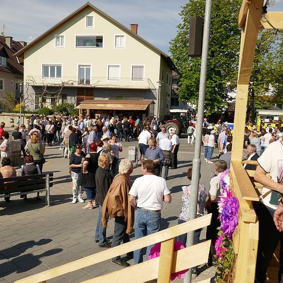 Eine Menschenmenge steht draußen, einige in einer Schlange, andere unterhalten sich, auf einem Stadtplatz mit Gebäuden und einem Traktor im Hintergrund.