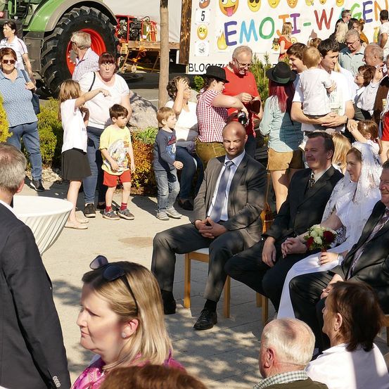 Eine Hochzeitszeremonie findet mit einer großen Gruppe von Menschen statt, einschließlich des Bräutigams und der Braut, die um sie herum stehen und sitzen. Ein Banner mit Emojis ist im Hintergrund.