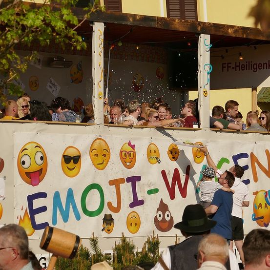 Menschen versammeln sich bei einer Veranstaltung im Freien und beobachten, wie eine Person Konfetti von einer Balkon wirft. Das Banner zeigt Emojis und das Wort EMOJI-WORLD.