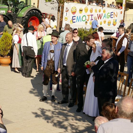 Eine Menschenmenge steht draußen, ein Mann in Anzug spricht in ein Mikrofon, eine Frau in weißem Kleid und andere in traditioneller bayerischer Kleidung. Ein Traktor ist im Hintergrund geparkt.