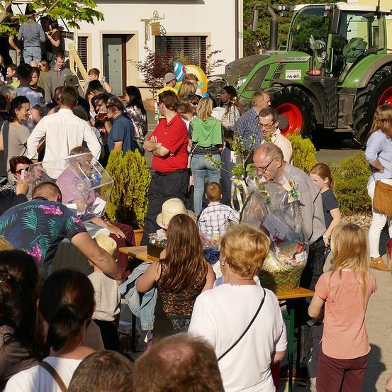 Eine Menschenmenge versammelt sich im Freien, einige sitzen und andere stehen, um Tische mit abgedeckten Gegenständen herum. Ein großer grüner Traktor ist in der Nähe geparkt und ein Gebäude ist im Hintergrund sichtbar.