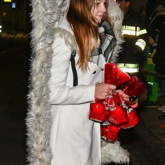Eine Frau in einem weißen Mantel mit Pelzbesatz hält rote Taschen in den Händen und steht in einem dunklen Bereich. Hinter ihr ist ein Feuerwehrmann in Uniform zu sehen.