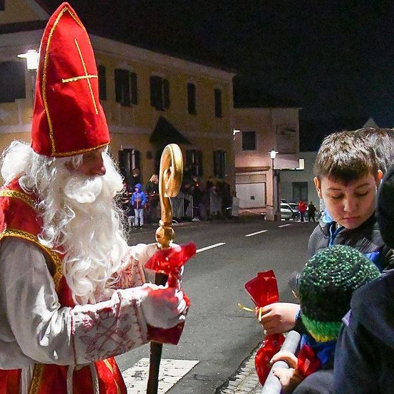 Ein Mann in der Kleidung des Heiligen Nikolaus verteilt Süßigkeiten an einen Jungen und ein Mädchen in einer nächtlichen Umgebung.