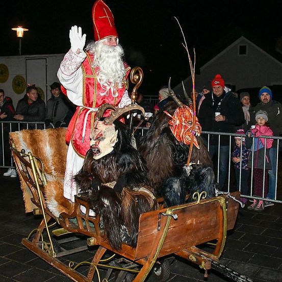 Ein Mann in Santa-Claus-Verkleidung winkt und reitet auf einem Schlitten mit zwei Ziegen, vor einer Menge bei Nacht auftretend.