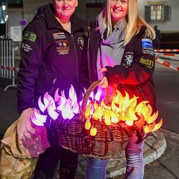 Zwei Frauen stehen draußen bei Nacht. Eine hält einen Korb mit leuchtenden, beleuchteten Früchten, während die andere eine Jacke mit einem Logo trägt. Hinter ihnen steht eine Metallabsperrung.