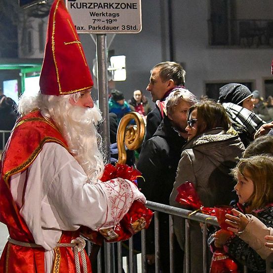 Eine Person als Weihnachtsmann verteilt rote Tüten an eine Menschenmenge auf der Straße. Das Schild zeigt Kurzparkzone und 7:00 bis 19:00 Uhr an.