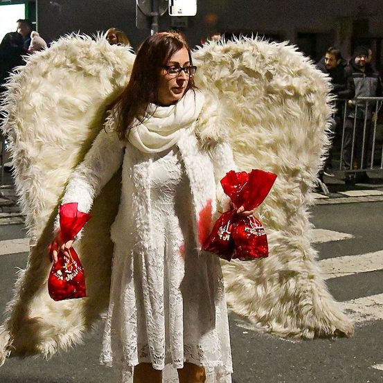 Eine Frau in einem Engelskostüm steht mit Flügeln auf der Straße. Sie trägt ein weißes Kleid und eine Brille. Sie hält zwei rote Taschen. Hinter ihr stehen Leute und eine Metallbarriere.