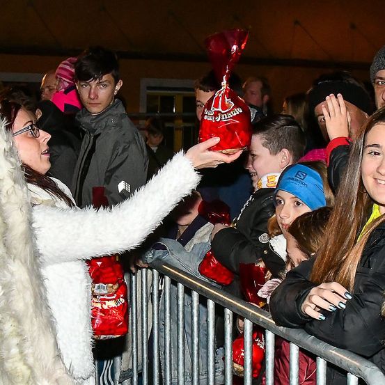 Eine Frau verteilt rote Süßigkeitentüten an Kinder in einer Menschenmenge, einige in Winterkleidung und Mützen. Die Kinder greifen eifrig nach den Tüten.