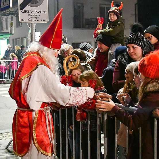 Eine Feier zum Sankt-Nikolaus-Tag, bei der ein Mann als Weihnachtsmann verkleidet Geschenke an eine Menschenmenge aus Kindern und Erwachsenen hinter einem Metallzaun verteilt.