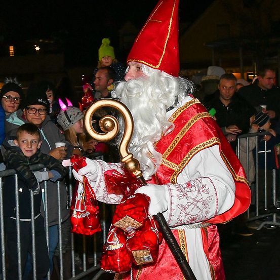 Ein Mann in Santa-Claus-Verkleidung verteilt Süßigkeiten an Kinder und hält einen Stab in der Hand, umgeben von einer Menschenmenge hinter einem Zaun.