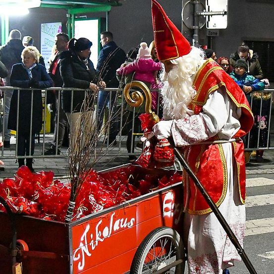 Eine als Sankt Nikolaus verkleidete Person verteilt Geschenke aus einem roten Karren, viele Menschen beobachten.