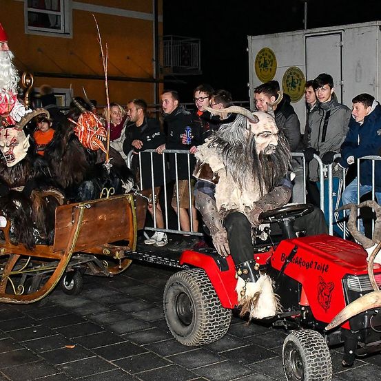 Eine Person in einem hörnertragenden Kostüm fährt einen roten Traktor mit angehängtem Schlitten, während Zuschauer hinter einem Metallzaun zuschauen.