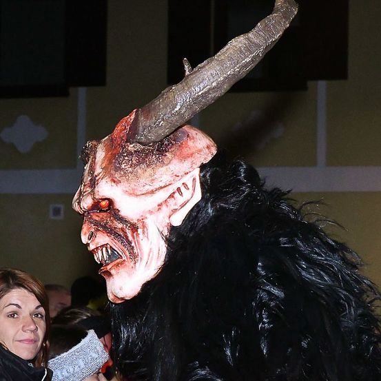 Eine Person in einem Kostüm mit einer teuflischen Maske steht vor einer Menge. Die Maske hat Hörner und rote Augen.