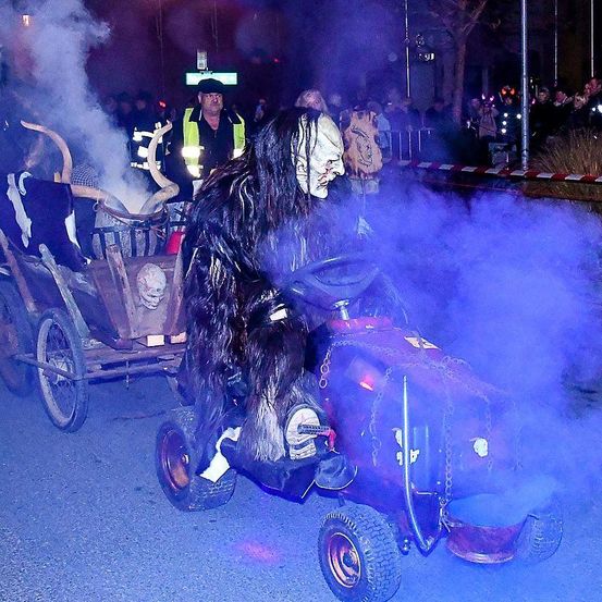 Eine Person in einem Monsterkostüm fährt einen Spielzeugtraktor mit Raucheffekten, gefolgt von einem Wagen mit Hörnern und einer Menge zuschauender Menschen.