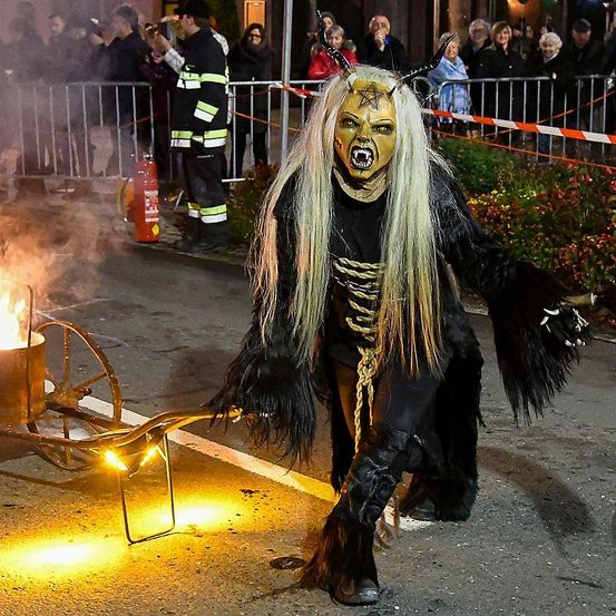 Eine Person mit einer gehörnten Maske und einer langen Perücke, die einen brennenden Wagen auf der Straße zieht. Ein Feuerlöscher ist in der Nähe, und eine Menschenmenge schaut zu.