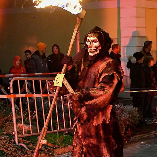 Eine Person, die als Sensenmann verkleidet ist, hält eine flammende Sense. Die Zahl sechs ist auf der Sense. Hinter der Person stehen mehrere Menschen, die möglicherweise eine Halloween-Veranstaltung beobachten.