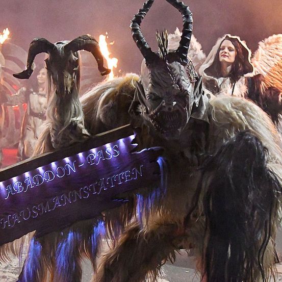 Eine Person verkleidet als Monster mit Hörnern und einem Schild, das 'Abaddon-Pass' besagt. Im Hintergrund halten Personen Fackeln und sind in Kostümen gekleidet.