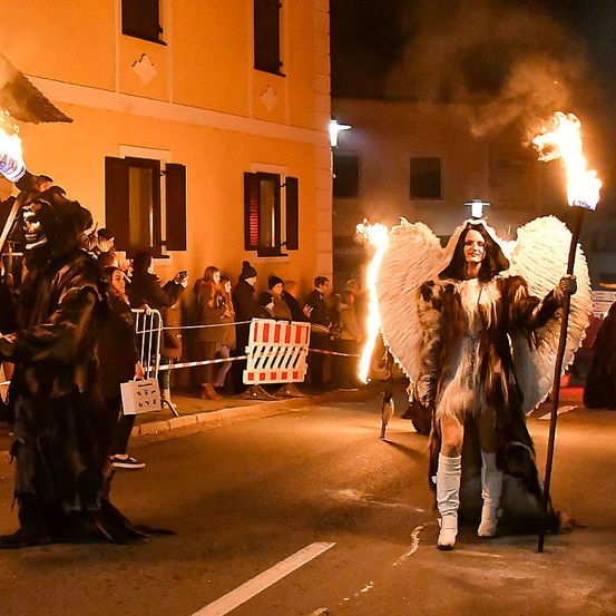 Zwei Personen in Kostümen und mit Fackeln gehen bei Nacht auf der Straße, während eine Menge zuschaut.