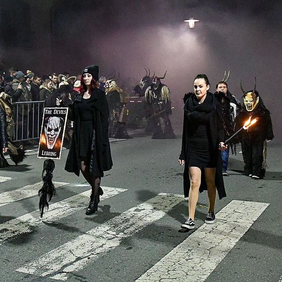 Zwei Frauen in Schwarz, eine mit einem Schild und die andere mit einer Fackel, überqueren eine Straße mit weißen Markierungen. Dahinter marschieren mehrere Personen in Hörnerkostümen. Ein Publikum beobachtet sie hinter einer Metallbarriere. Die Szene wird von einer Straßenlampe beleuchtet.