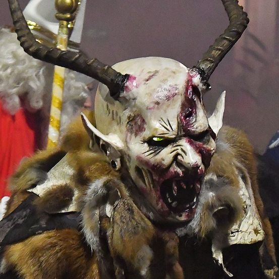 Ein Nahaufnahme eines Menschen, der eine Hörner-Dämonenmaske mit blutigem und wütendem Gesicht trägt. Die Maske ist mit künstlichem Blut bedeckt und hat eine fellartige Textur.