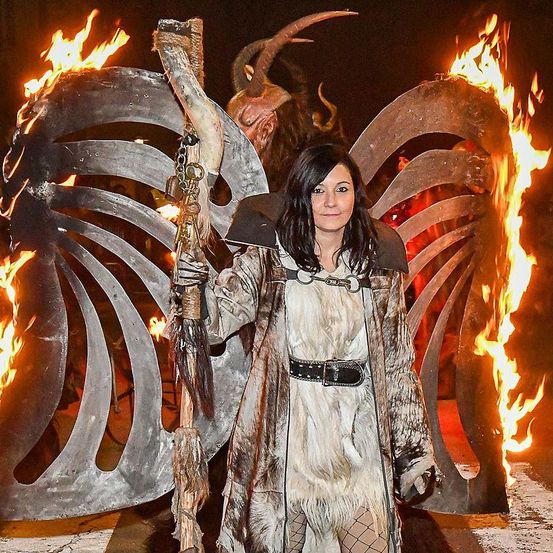 Eine Frau in einem Pelzmantel und einer gehörnten Maske posiert nachts vor einem brennenden Feuerwerk für ein Foto.