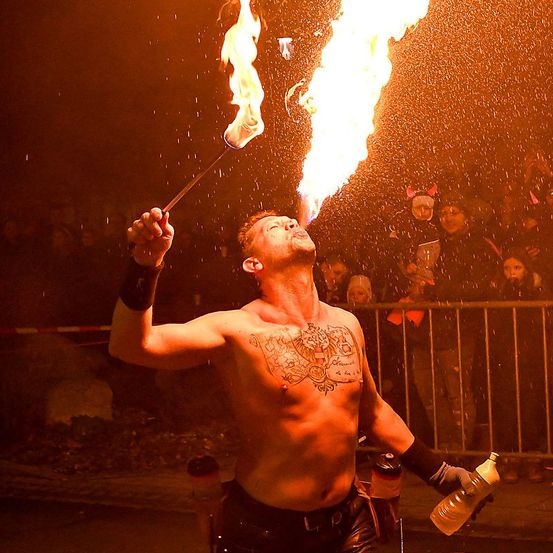 Ein tätowierter Mann ohne Oberbekleidung bläst Feuer aus einem Stock und führt es vor einem Publikum vor. Das Publikum beobachtet ihn hinter einem Zaun.