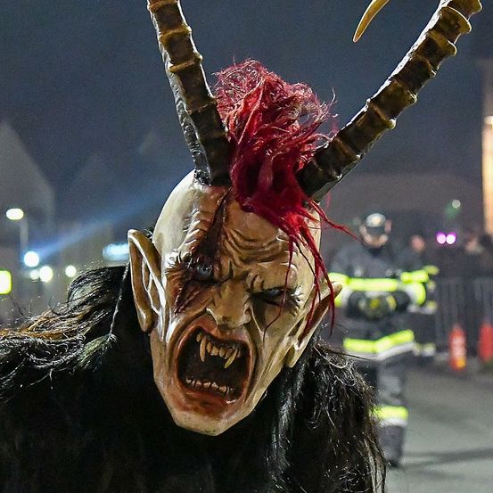 Eine Person mit einer gehörnten Maske steht im Vordergrund mit einer Menschenmenge und Feuerwehrleuten im Hintergrund.