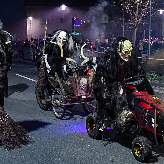 Ein Halloween-Umzug mit Menschen in gruseligen Kostümen auf kleinen Traktoren. Eine Person fährt und eine andere zieht einen Wagen mit einer Besen.
