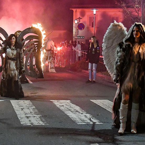 Menschen in Kostümen, darunter Engelsflügel, stehen auf einer Straße mit einem Feuer und einer Menschenmenge im Hintergrund.