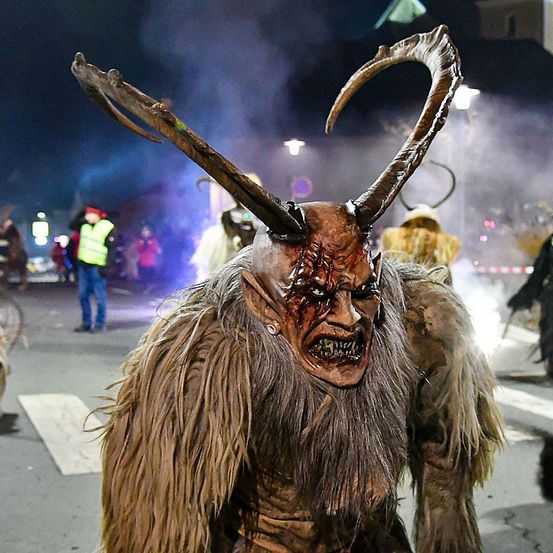 Eine Person in einem Kostüm mit großen Hörnern steht bei Nacht auf der Straße, mit Rauch und anderen Leuten um sie herum.