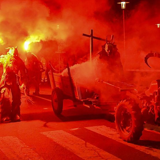 In der Nacht stehen Menschen in Kostümen vor einem rot beleuchteten Traktor mit Rauch und einem Kreuz im Hintergrund.