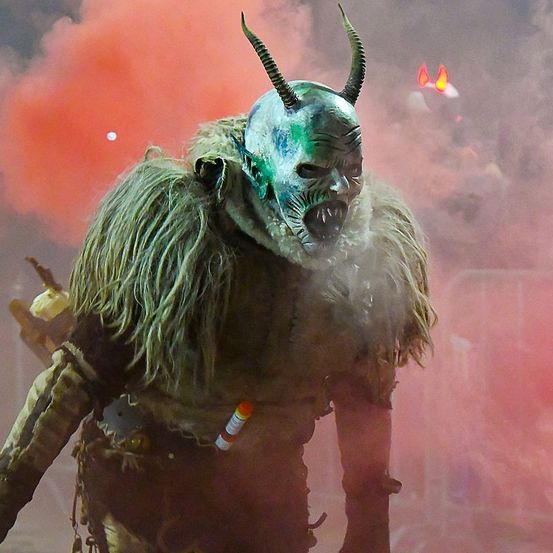Eine Person in einer gehörnten Maske steht in einem rauchgefüllten Bereich, wahrscheinlich bei einem Auftritt. Die Maske hat grüne und weiße Farben, und der Rauch ist pink.