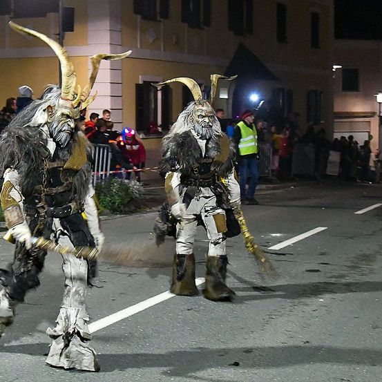 Zwei in Teufelskostüme gekleidete Personen gehen bei Nacht eine Straße entlang, umgeben von einer Menge Zuschauer.