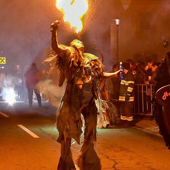 Ein Performer bläst Feuer aus dem Mund während einer nächtlichen Veranstaltung, mit Zuschauern und einer Barriere in der Nähe.
