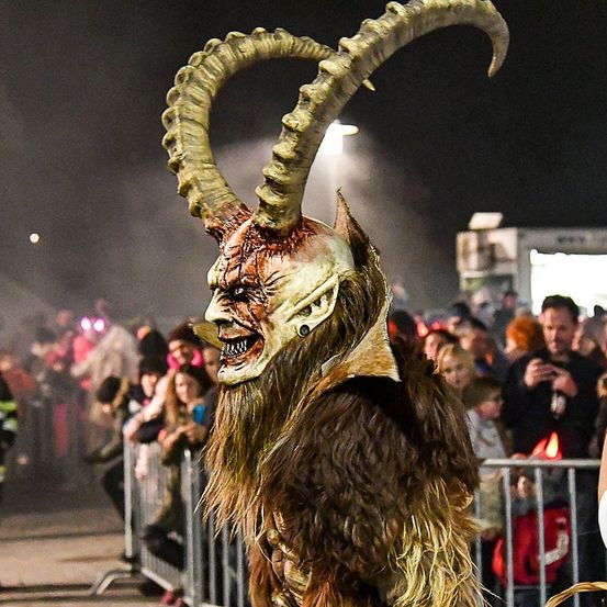 Eine Person in einem Hörnerkostüm steht vor einer Menschenmenge bei Nacht. Die Menge steht hinter einer Metallbarriere. Einige Leute halten Getränke in den Händen.