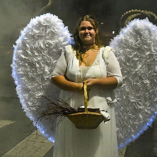Eine Frau in einem weißen Engelkostüm mit großen weißen Engelflügeln am Rücken hält einen Korb mit einigen Büchern darin.