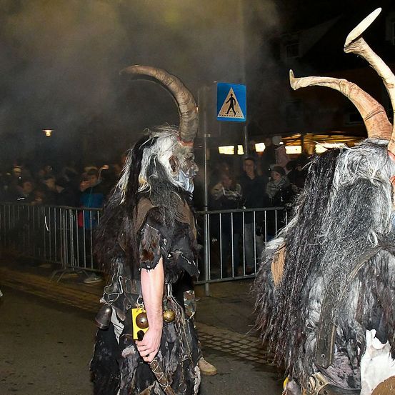 Zwei Personen in Hörnerkostümen gehen bei Nacht vor einer Menge. Sie sind von einem Metallzaun umgeben, und ein Fußgängerüberweg-Schild steht hinter ihnen.