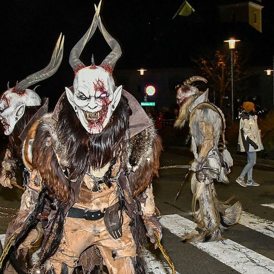 Drei Menschen mit Hörnermasken laufen nachts auf einer Straße. Sie gehören zu einer Gruppe, die Tierkostüme trägt. Ein Fußgänger geht auf dem Bürgersteig vorbei.