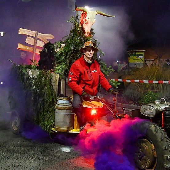 Ein Mann in einer roten Jacke fährt einen dekorierten Traktor mit einem Kuhschädel, der lila Rauch abgibt. Der Traktor ist von Grün umgeben.