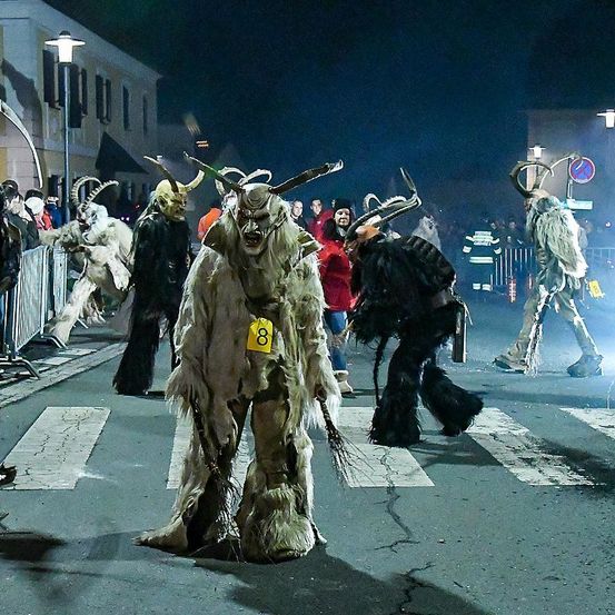 Eine Gruppe von Menschen in Hörnerkostümen läuft nachts eine Stadtstraße entlang, mit einer Menge Zuschauern und Straßenlaternen im Hintergrund.