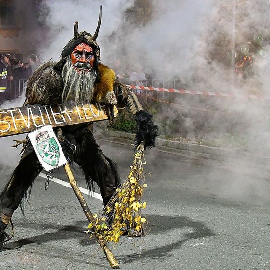 Eine Person in einem gehörnten Kostüm hält ein Holzschild mit der Aufschrift 'St.Villier-Feuer'. Sie hat einen Bart und einen Stab und steht auf einer Straße mit Rauch drumherum.