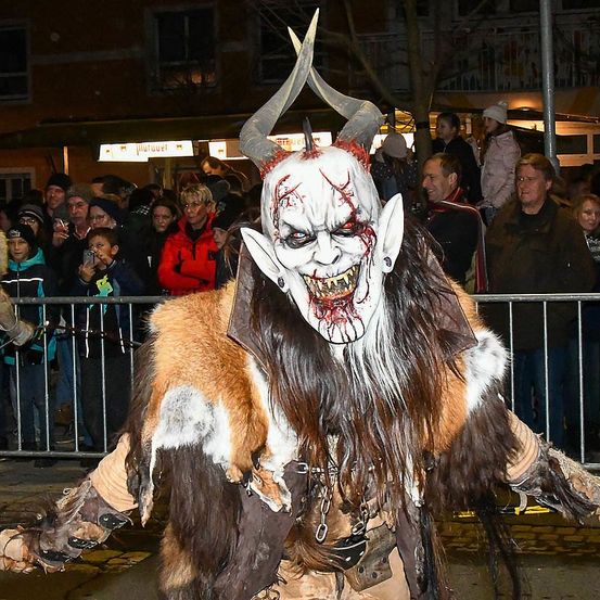 Eine Person in einem Monsterkostüm mit Hörnern und künstlichem Blut im Gesicht steht vor einer Menschenmenge. Die Menge beobachtet hinter einem Metallzaun.