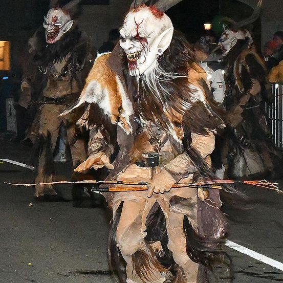 Eine Person in einem gruseligen Monsterkostüm geht nachts auf der Straße und hält einen langen Bogen. Hinter ihnen sind mehrere Personen ebenfalls in ähnlichen Kostümen. Die Straße ist dunkel, nur wenige Lichter erhellen den Bereich.