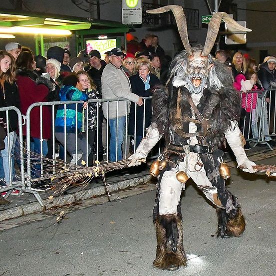 Eine Person in einem gehörnten Kostüm geht mit einem Besen, während die Leute hinter einem Zaun zusehen. Die Menge ist vielfältig und trägt Winterkleidung. Die Umgebung scheint ein Outdoor-Event bei Nacht zu sein.