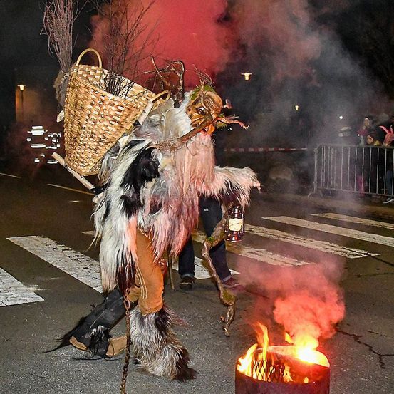 Eine Person in einem Kostüm steht bei Nacht auf der Straße, hält eine Laterne und einen Korb mit Zweigen und steht neben einem Feuer. Eine Menschenmenge beobachtet.