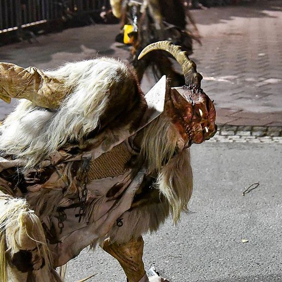 Eine Nahaufnahme einer kostümierten Person, die einem Dämon oder Ziegenbock mit Hörnern ähnelt, auf einem gepflasterten Straßenabschnitt.