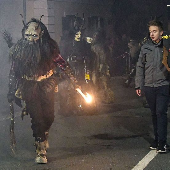 Ein Mann in einem Monsterkostüm mit einer brennenden Fackel geht auf der Straße, mit einer anderen Person in einem Monsterkostüm hinter ihm. Sie sind von einer Menschenmenge in einem dunklen Bereich umgeben.