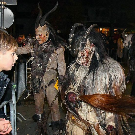 Ein Kind beobachtet zwei gehörnte Kostümfiguren im Freien bei Nacht, mit einer Menge und Gebäuden im Hintergrund.